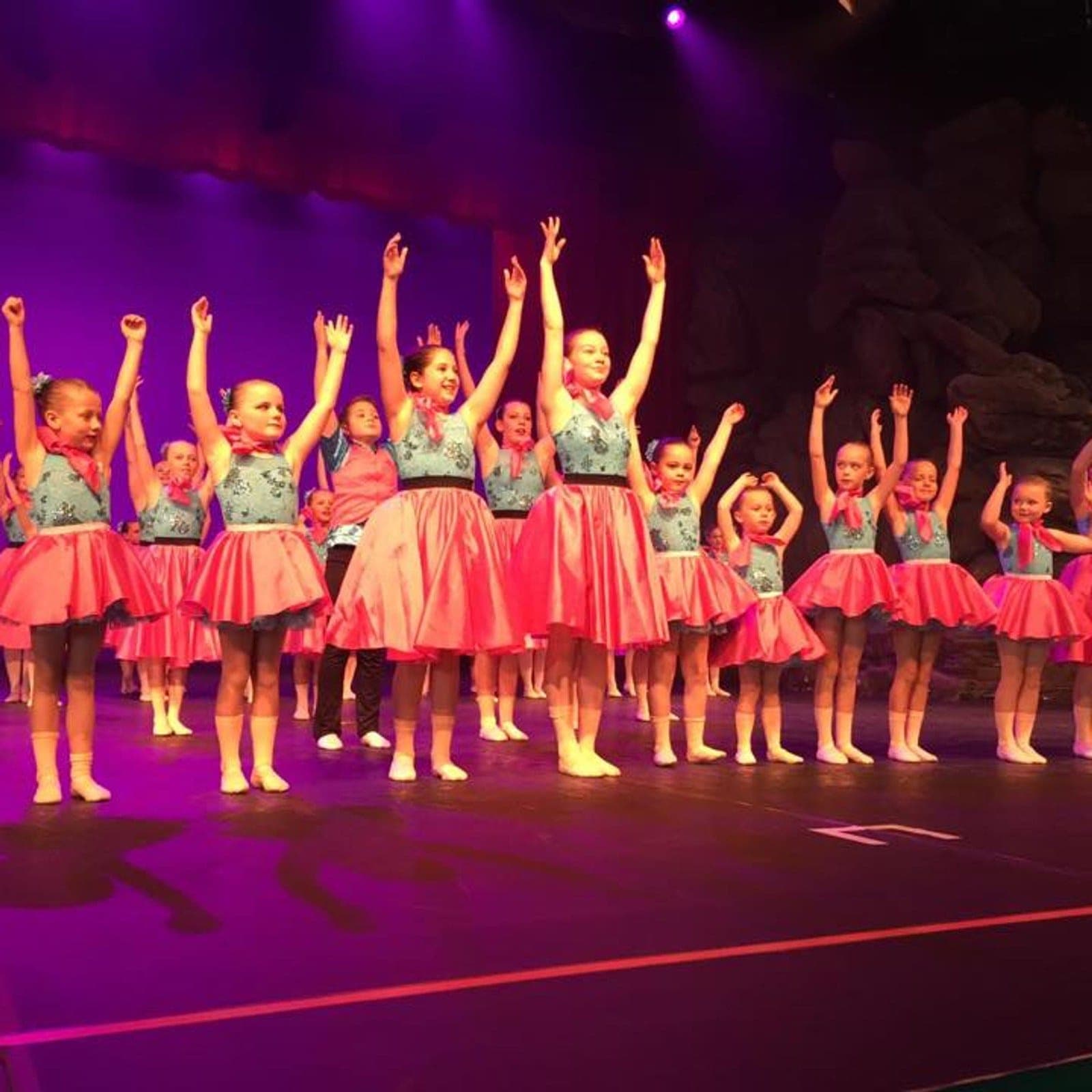 Group of dancers in blue bodices and magenta skirts under purple stage light
